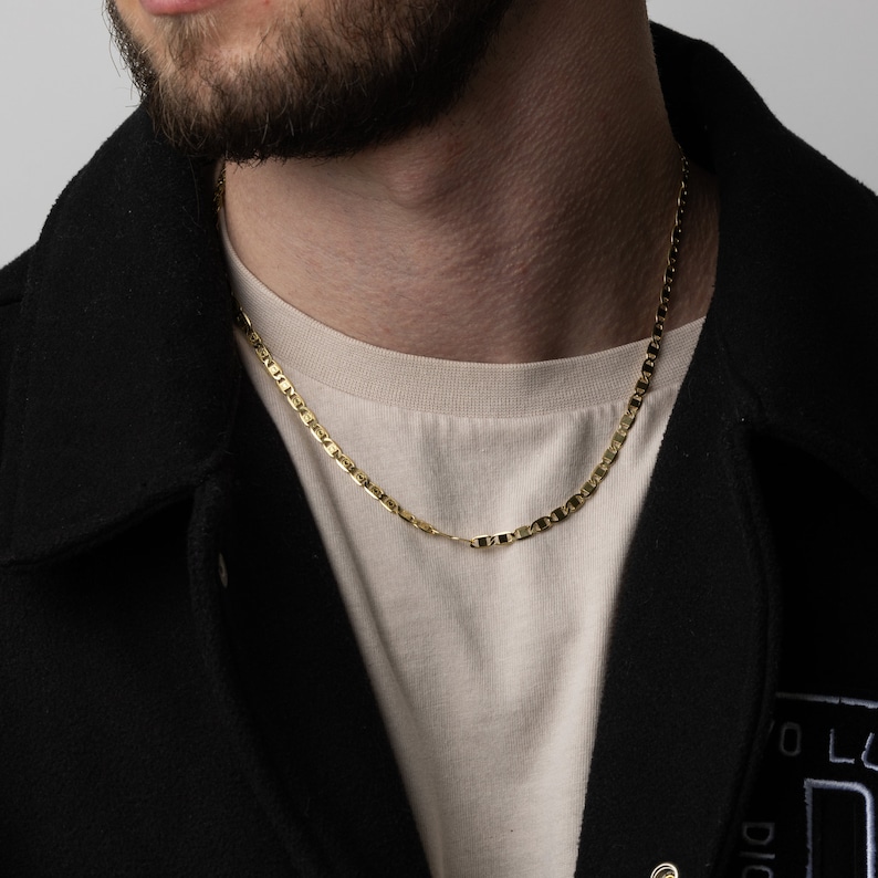 May include: A gold chain necklace with a mariner link design. The chain is worn by a person wearing a black jacket and a beige t-shirt.