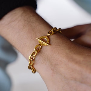 May include: A gold-colored chain bracelet with a toggle clasp. The bracelet is made of thick, rounded links and features a circular clasp with a bar closure. The bracelet is worn on a wrist.