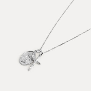 May include: A silver-colored necklace featuring two pendants: an oval pendant with a religious figure and a crucifix. The necklace has a delicate chain and is set against a white background. The pendants are detailed and shiny.