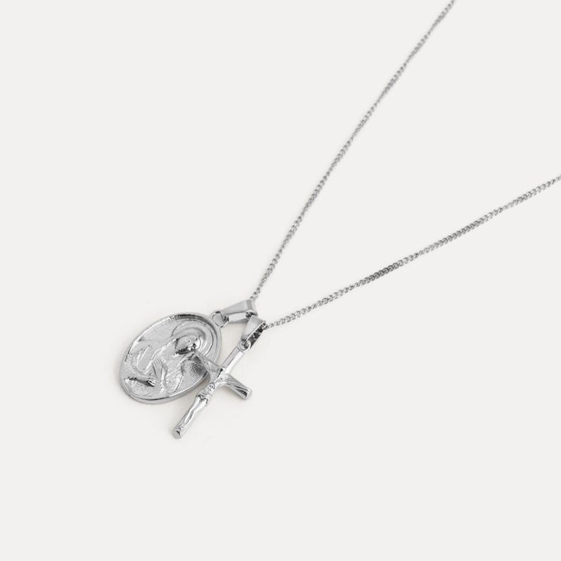 May include: A silver-colored necklace featuring two pendants: an oval pendant with a religious figure and a crucifix. The necklace has a delicate chain and is set against a white background. The pendants are detailed and shiny.