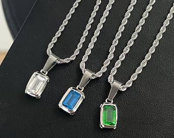 May include: Three silver-toned necklaces with rectangular gemstone pendants in clear, blue, and green. The pendants are set in a silver bezel and hang from a rope chain. The necklaces are displayed on a black surface.