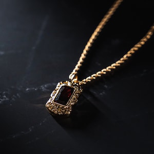 May include: A gold-colored necklace with a rectangular, dark red gemstone pendant. The pendant is framed by an ornate, filigree design. The chain is a twisted rope style. The background is a dark, textured surface.