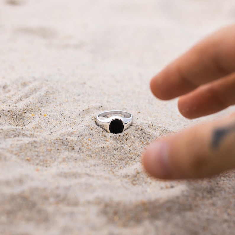 May include: A silver ring with a black oval stone set in the center. The ring is lying on a bed of white sand.