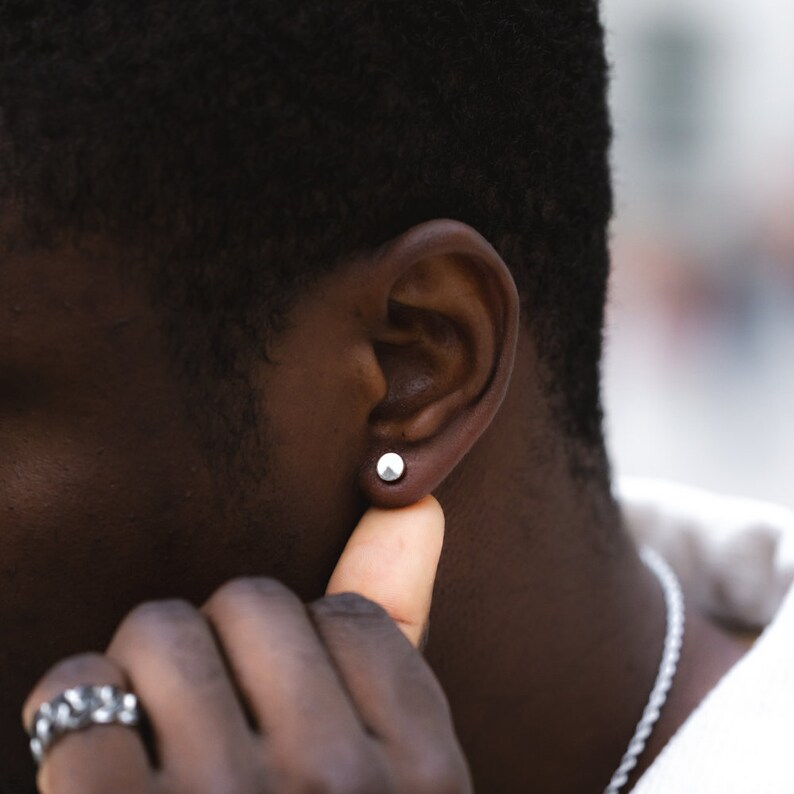 May include: A close-up of a person's ear with a small, round, silver earring. The person is wearing a white shirt and has short black hair.