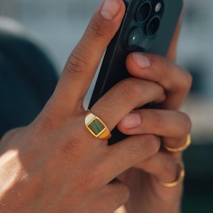 Könnte beinhalten: Ein goldener Siegelring mit einem rechteckigen grünen Stein und weißen Akzenten. Der Ring befindet sich an einem Finger, mit einem schwarzen Telefon im Hintergrund. Ein weiterer goldener Ring ist ebenfalls sichtbar.