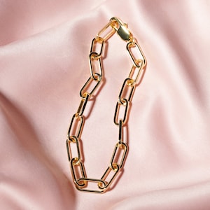 May include: A gold-colored chain bracelet with elongated rectangular links. The bracelet is displayed on a pink satin surface, with the clasp visible at the top. The jewelry is a classic accessory.