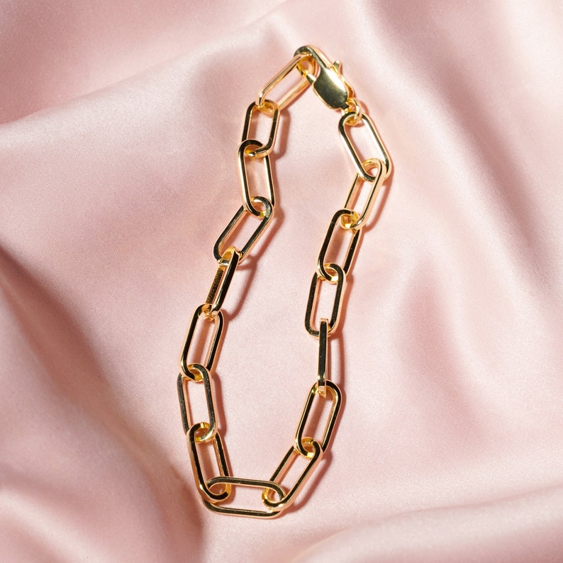 May include: A gold-colored chain bracelet with elongated rectangular links. The bracelet is displayed on a pink satin surface, with the clasp visible at the top. The jewelry is a classic accessory.