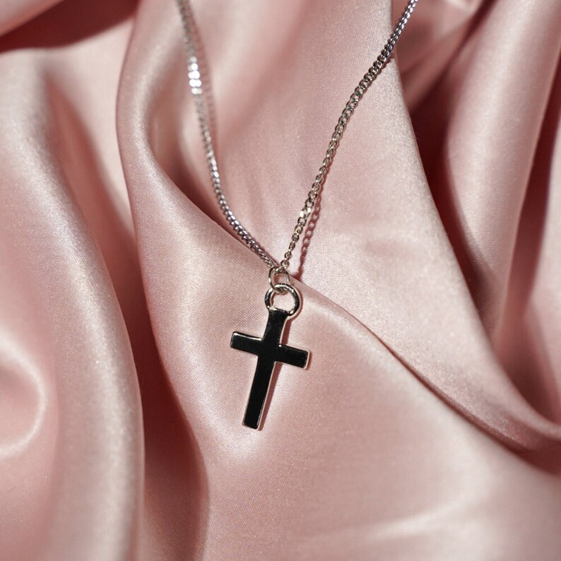 May include: A silver chain necklace with a simple silver cross pendant. The necklace is laying on a pink satin fabric.