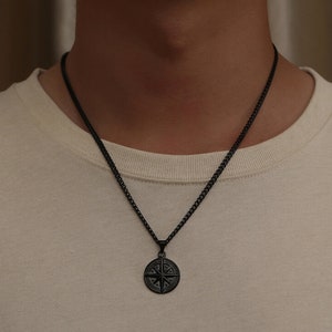 K&ouml;nnte beinhalten: Eine schwarze Kettenkette mit einem schwarzen Kompass-Anh&auml;nger. Der Kompass hat ein detailliertes Design mit Himmelsrichtungen.