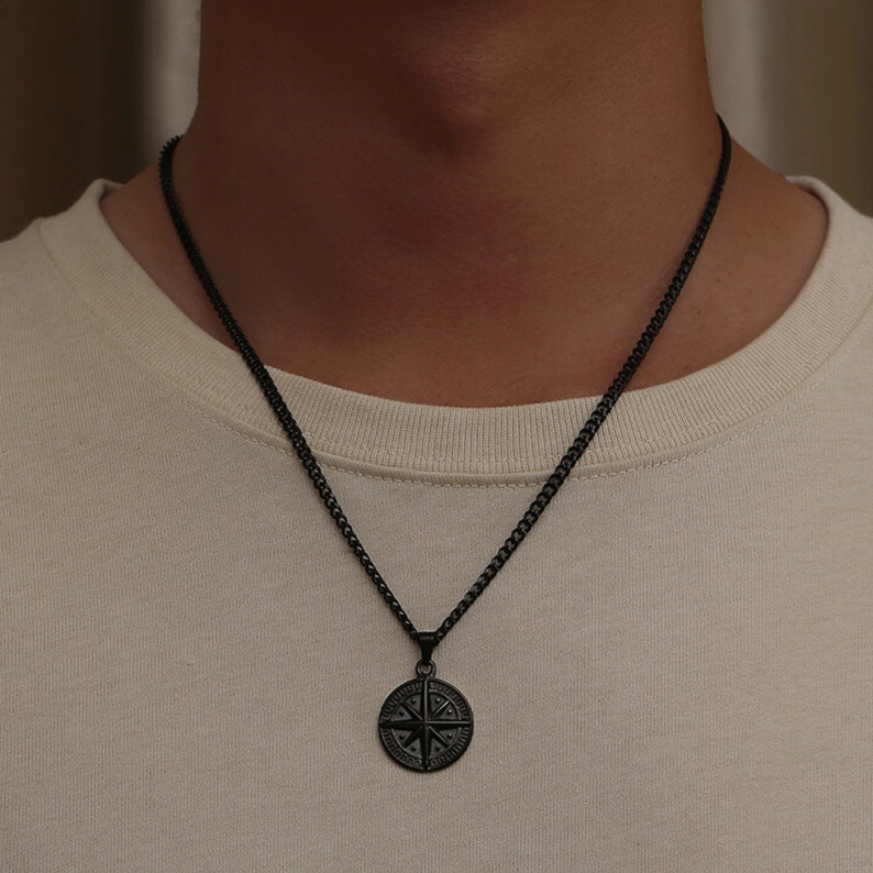 K&ouml;nnte beinhalten: Eine schwarze Kettenkette mit einem schwarzen Kompass-Anh&auml;nger. Der Kompass hat ein detailliertes Design mit Himmelsrichtungen.