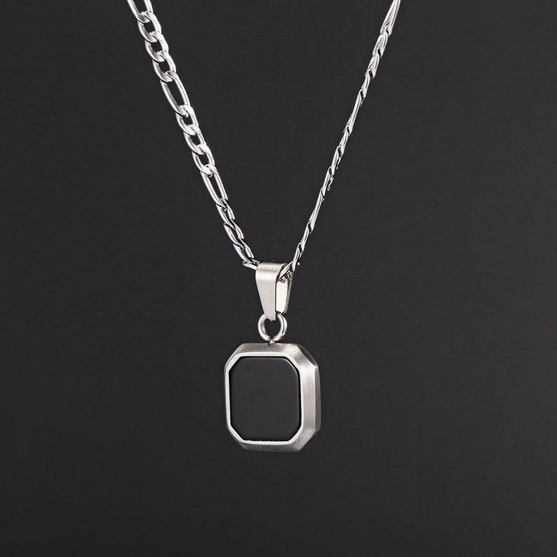 May include: A silver necklace with a geometric pendant. The pendant has a black square center and a silver frame. The necklace chain is a combination of a curb chain and a thinner chain.
