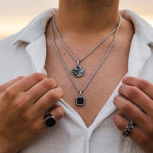 May include: Two silver necklaces with pendants, one round with a dragon design and one square with a black center. The necklaces are layered and worn over a white shirt. A black ring is visible on the left hand.