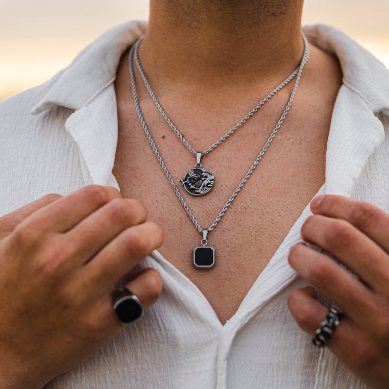 May include: Two silver necklaces with pendants, one round with a dragon design and one square with a black center. The necklaces are layered and worn over a white shirt. A black ring is visible on the left hand.