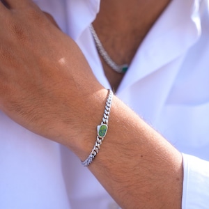 May include: A silver curb chain bracelet with a rectangular green stone set in the center. The bracelet is worn on a person's wrist, against a white shirt. The bracelet's links are closely spaced, creating a textured appearance.
