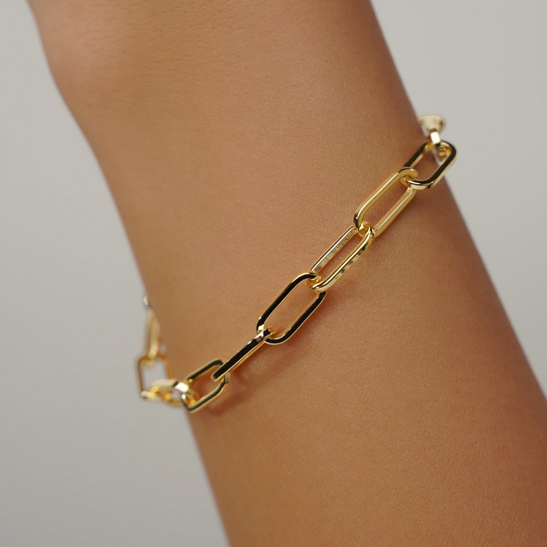 May include: A gold-colored chain bracelet with rectangular links. The bracelet is worn on a person's arm, and the links are connected in a repeating pattern. The bracelet has a shiny finish and is a simple, elegant piece of jewelry.