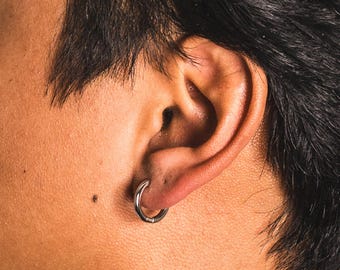 May include: Close-up of a silver hoop earring in a pierced ear. The earring is a simple, circular design. The skin around the ear is tan with dark hair in the background.