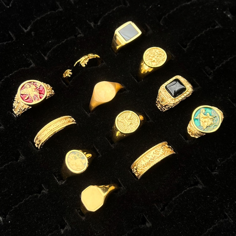 May include: A collection of eleven gold-toned rings arranged in a grid. The rings feature various designs, including solid bands, signet rings with black inlays, and circular faces with embossed figures in red, green, and gold metallic finishes.
