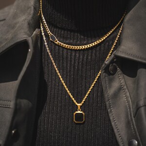 May include: Gold-toned necklaces layered on a black turtleneck. The top necklace has a black oval accent, while the lower necklace features a square black pendant. The necklaces are set against a black jacket.