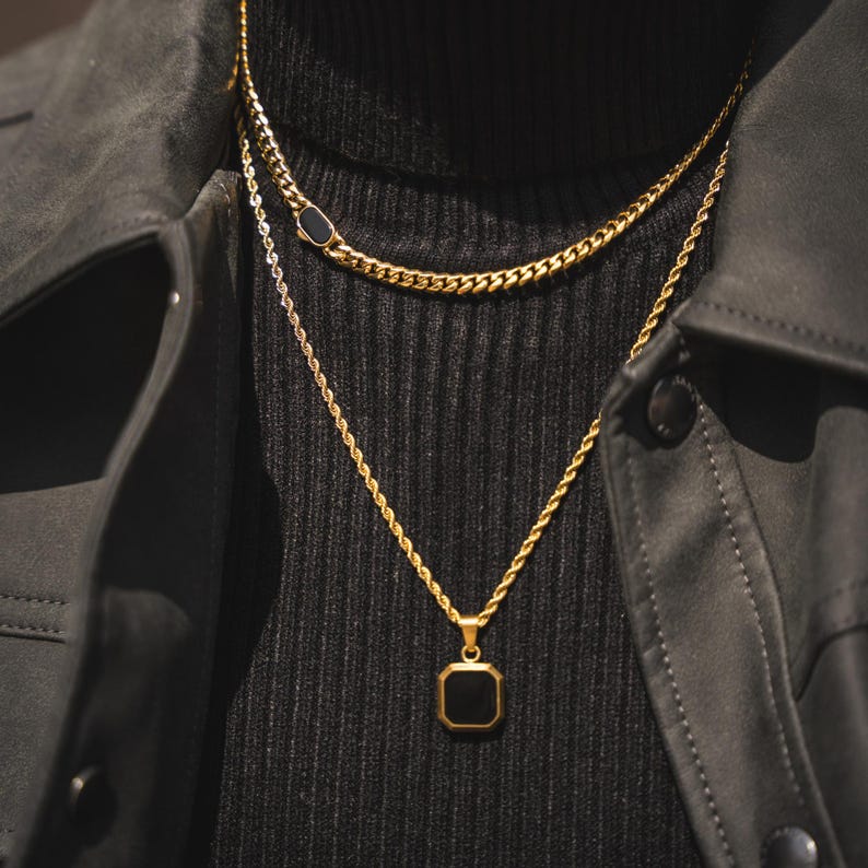 May include: Gold-toned necklaces layered on a black turtleneck. The top necklace has a black oval accent, while the lower necklace features a square black pendant. The necklaces are set against a black jacket.