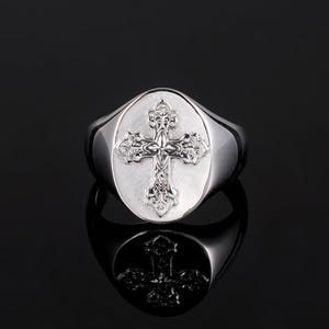 May include: A silver signet ring with an oval face featuring a detailed cross design.