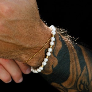 Puede incluir: Un brazalete de perlas blancas con detalles dorados en la muñeca de una persona.
