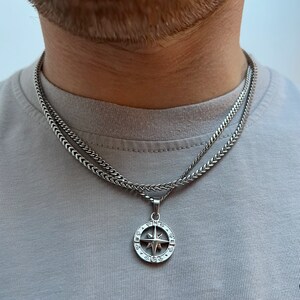 May include: A silver-colored necklace with a compass pendant. The pendant features a star design within a circular frame, suspended from a wheat chain. The necklace is layered over a second, similar chain. The person is wearing a gray t-shirt.