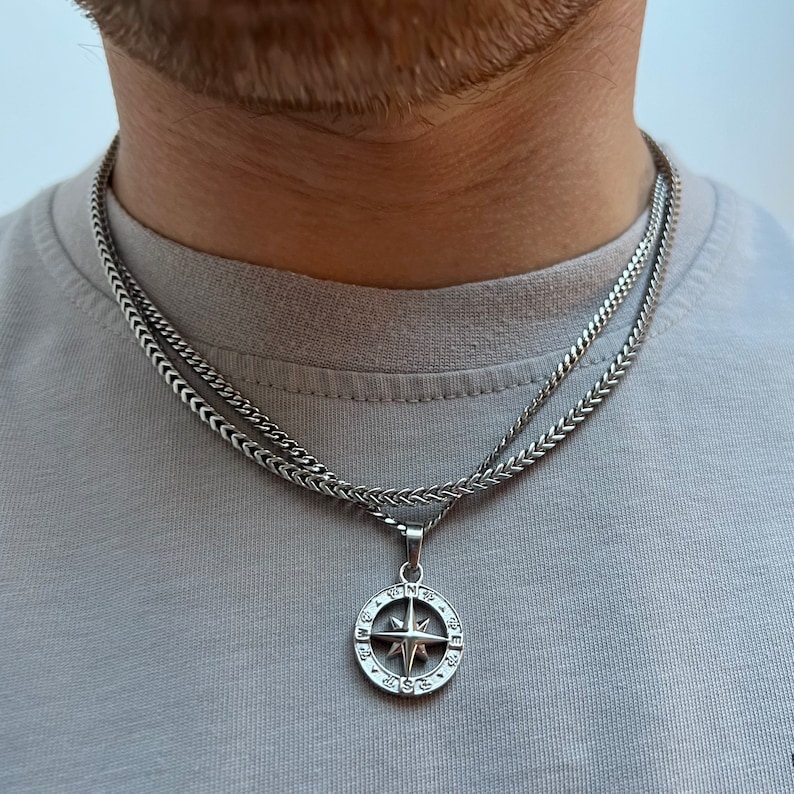 May include: A silver-colored necklace with a compass pendant. The pendant features a star design within a circular frame, suspended from a wheat chain. The necklace is layered over a second, similar chain. The person is wearing a gray t-shirt.