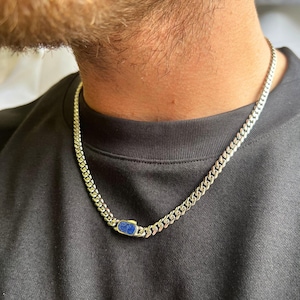 Peut inclure: Un collier chaîne gourmette argenté avec un pendentif rectangulaire en pierre bleue. Le collier est porté sur un t-shirt noir. Les maillons de la chaîne sont rapprochés, créant un look audacieux et moderne. La pierre bleue ajoute une touche de couleur.