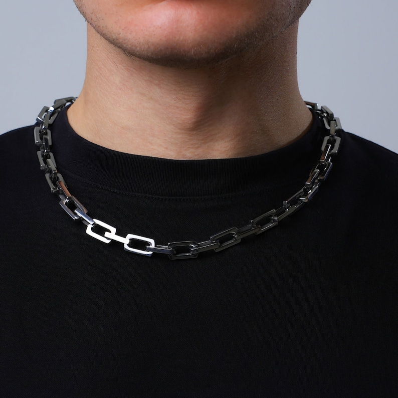 May include: A silver-colored chain necklace with rectangular links. The necklace is worn against a black shirt, creating a contrast. The chain is a bold accessory, suitable for various styles.