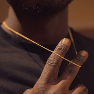 Könnte beinhalten: Nahaufnahme einer Hand, die eine goldene Kettenkette hält. Die Kette ist dünn und zart, und die Hand liegt auf der Brust.