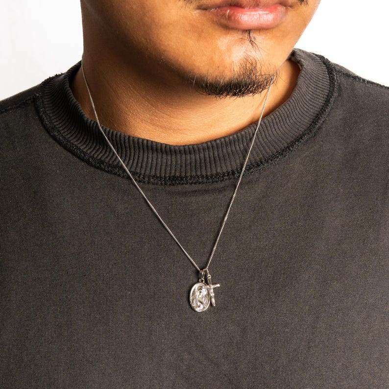 May include: Silver necklace with a pendant featuring a religious icon and a small cross. The necklace is worn against a dark gray crew neck shirt. The pendant has intricate detailing and hangs from a delicate chain.
