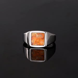 Puede incluir: Un anillo de sello plateado con una piedra cuadrada con un patrón naranja y marrón. El anillo está sobre un fondo oscuro, con un reflejo visible debajo. El diseño del anillo es simple y clásico.