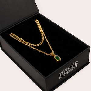 May include: A gold-toned layered chain necklace featuring a rectangular emerald-green pendant and a smaller matching green accent stone, presented in a black box with the text TWISTED PENDANT printed in white on the interior base of the box.