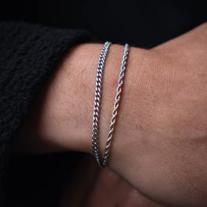 May include: Two silver bracelets on a wrist. One is a curb chain, the other a rope chain. The bracelets are made of a shiny, metallic material. The background is a dark, solid colour.