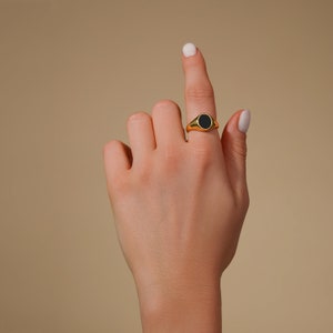 Op de afbeelding: Een gouden zegelring met een zwarte onyx steen. De ring heeft een gepolijste, glanzende afwerking. De ring wordt getoond op een hand tegen een neutrale achtergrond.