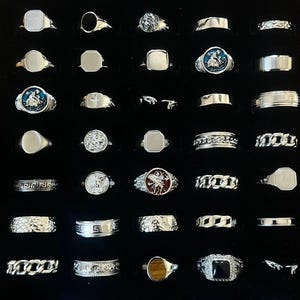 May include: A collection of silver-toned rings arranged in a grid, featuring various designs including signet rings, chain-link bands, textured surfaces, and rings with inlaid stones or circular emblems depicting figures and symbols in blue and red.