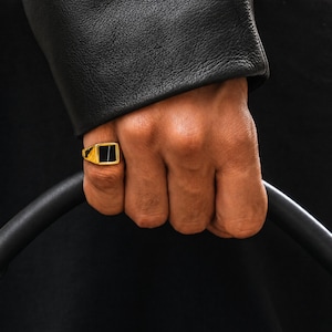 May include: A gold-colored signet ring with a black rectangular stone. The ring is on a finger, with a black leather jacket in the background. The ring is a classic piece of jewelry.