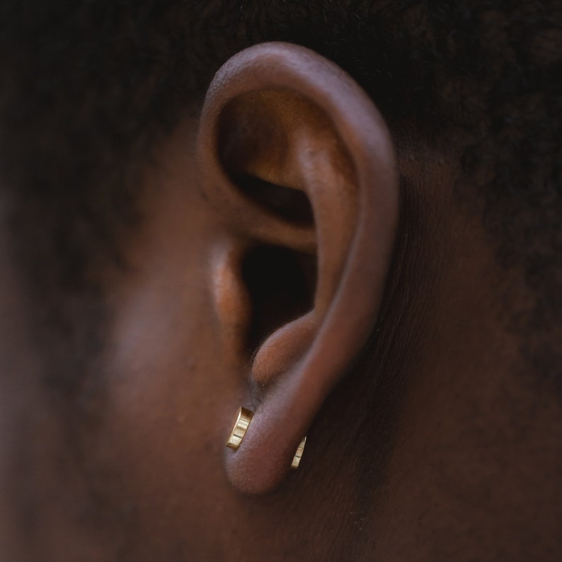 May include: A close-up of a person's ear with two small gold hoop earrings. The earrings are simple and understated, and they add a touch of elegance to the person's look.