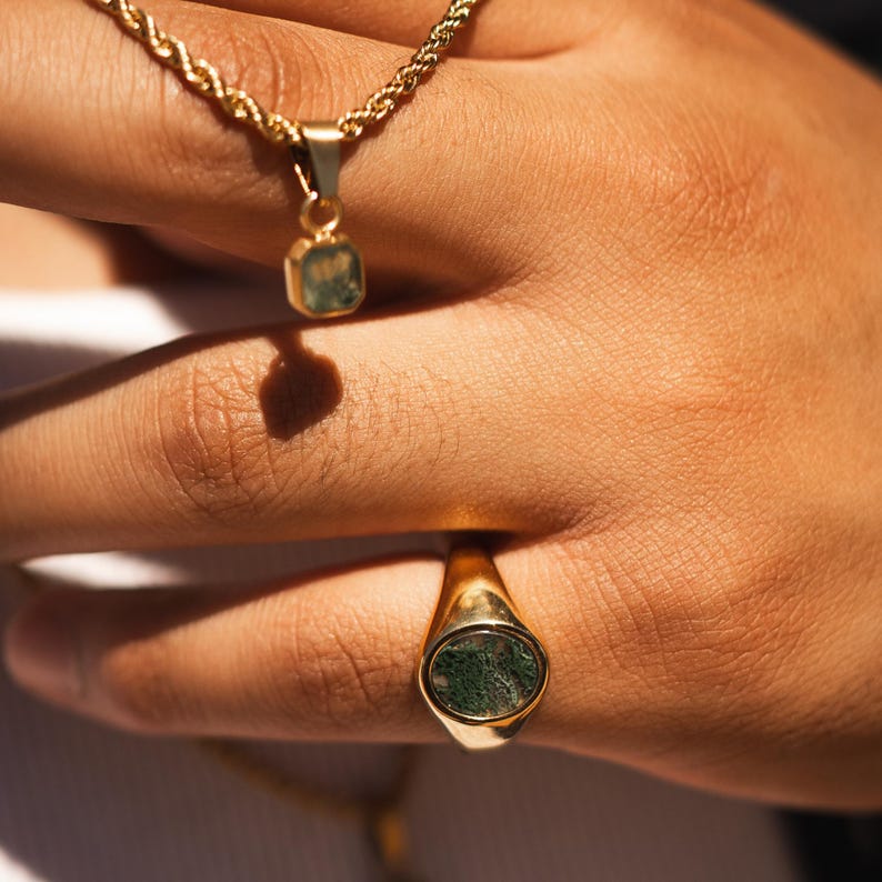 May include: Gold-toned jewelry set featuring a necklace with a square pendant and a signet ring. Both pieces have a green stone inlay. The necklace has a rope chain, and the ring is oval-shaped.