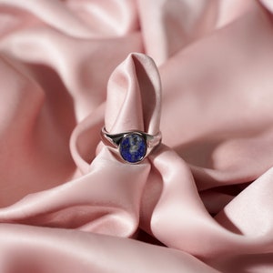 Op de afbeelding: Een zilveren ring met een blauwe lapis lazuli-steen gezet in een ovale vorm. De ring wordt getoond op een roze satijnen stof.