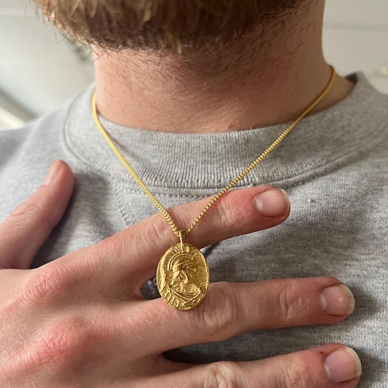 May include: A gold chain necklace with a gold pendant featuring a detailed profile of a person wearing a helmet.