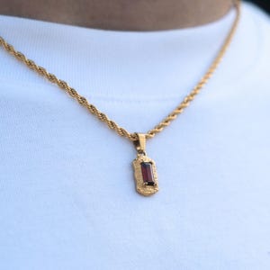 Op de afbeelding: Goudkleurige ketting met een rechthoekige, donkerrode edelsteen hanger. De hanger is gezet in een decoratief frame en hangt aan een touwketting. De ketting wordt getoond tegen een witte achtergrond.
