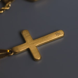 May include: A gold cross pendant with a brushed finish and a small engraved logo. The pendant is attached to a gold chain.