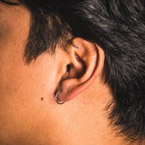 May include: Close-up of a person's ear with a silver hoop earring. The ear is set against a backdrop of dark hair and skin. The earring is a simple, circular design.
