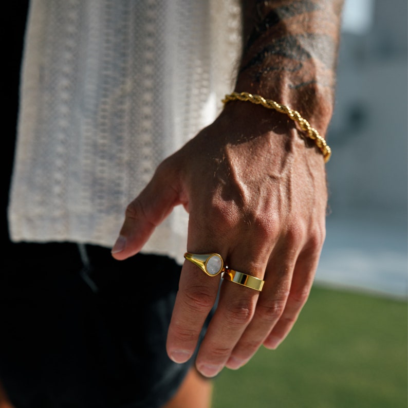 May include: A gold-colored bracelet and two rings are displayed on a hand. One ring has a white stone, and the other is a simple band. The bracelet and rings are made of a shiny, gold-colored metal. The hand is in front of a white, textured shirt.