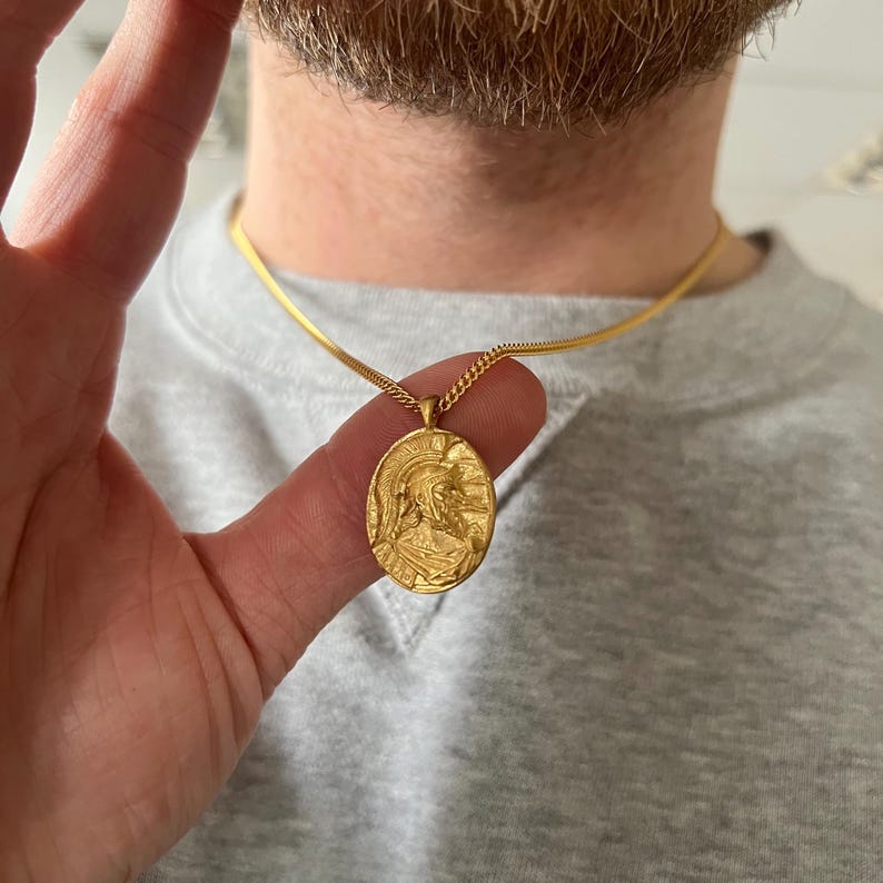 May include: A gold chain necklace with a gold pendant featuring a detailed profile of a person wearing a helmet.
