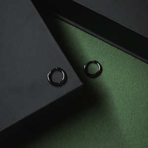 May include: A pair of black hoop earrings. The earrings are circular with a hinged clasp. The background is a dark grey surface and green fabric. The earrings are small and minimalist.