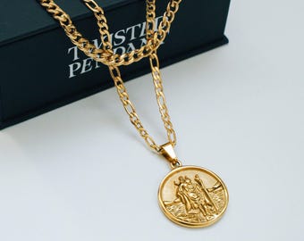 May include: Gold-colored necklace with a round pendant featuring a relief of two figures. The necklace has a Figaro chain design. The pendant is suspended from a small bail. The necklace is displayed on a black box with white text.