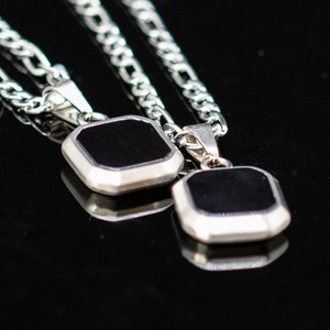 May include: Two silver necklaces with square, black pendants. The pendants have a silver border and are displayed on a reflective black surface. The chains are a classic curb link style.