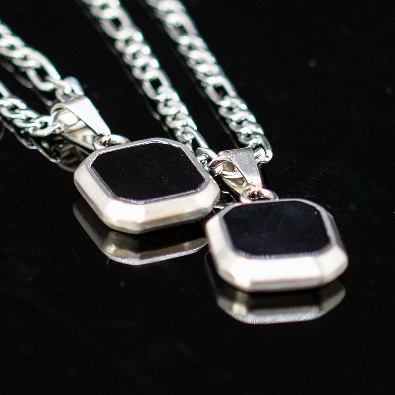 May include: Two silver necklaces with square, black pendants. The pendants have a silver border and are displayed on a reflective black surface. The chains are a classic curb link style.